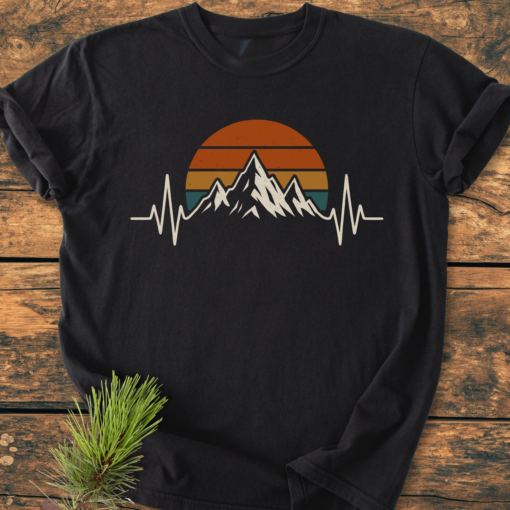 Mountain Heartbeat - Vintage Wash Tee: Black long-sleeve T-shirt with mountain-peak and EKG-line print, laid on wooden surface, accented by a pine sprig; embodies a hiker's passion and relaxed fit.