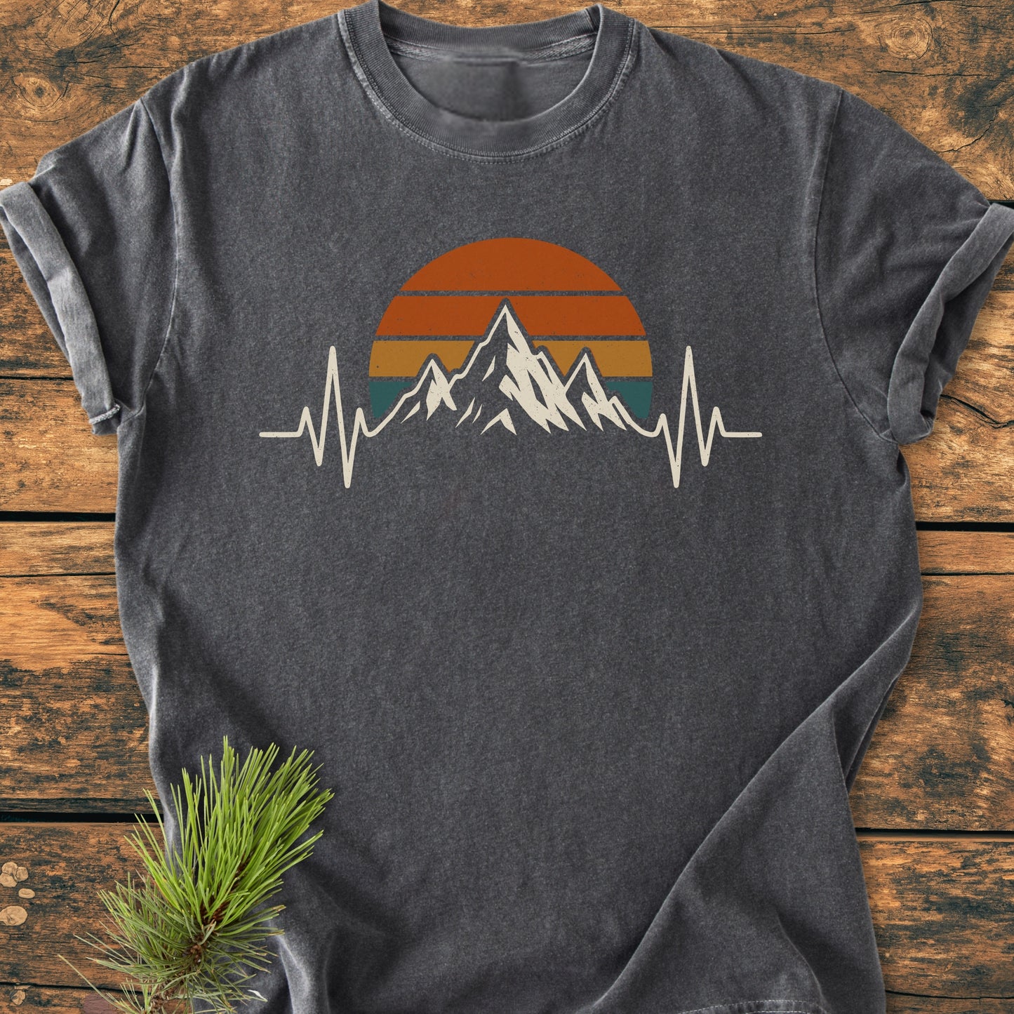 Mountain Heartbeat - Vintage Wash Tee featuring a mountain silhouette with heartbeat line, set against a retro sun motif. Displayed on a wooden surface with a pine branch accent.