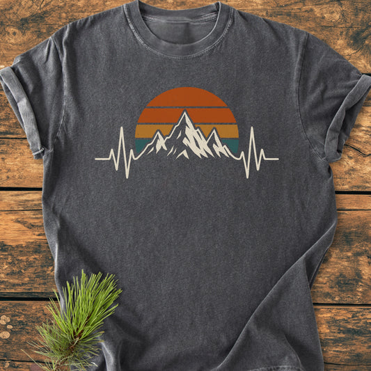 Mountain Heartbeat - Vintage Wash Tee featuring a mountain silhouette with heartbeat line, set against a retro sun motif. Displayed on a wooden surface with a pine branch accent.