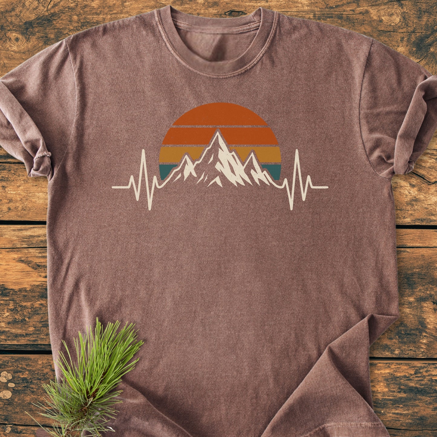 Mountain Heartbeat - Vintage Wash Tee: A brown-washed T-shirt featuring a sun, mountain, and ECG line graphic, laid flat on wood, showcasing its relaxed fit and vintage style, ideal for trail enthusiasts.