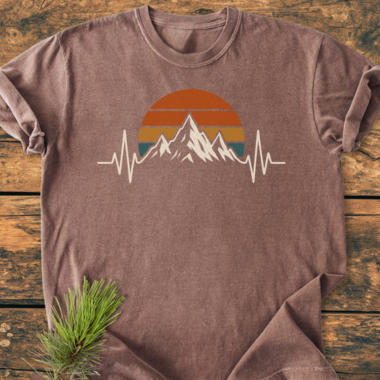 Mountain Heartbeat - Vintage Wash Tee: A brown-washed T-shirt featuring a sun, mountain, and ECG line graphic, laid flat on wood, showcasing its relaxed fit and vintage style, ideal for trail enthusiasts.