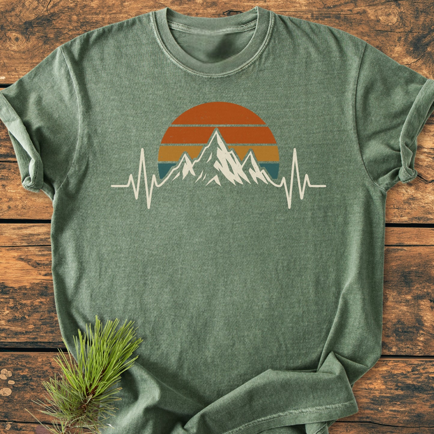 Mountain Heartbeat - Vintage Wash Tee displayed on a wooden surface, featuring a mountain and sunset graphic with an EKG line. A pine sprig adds a nature-inspired touch, highlighting its hiking theme.
