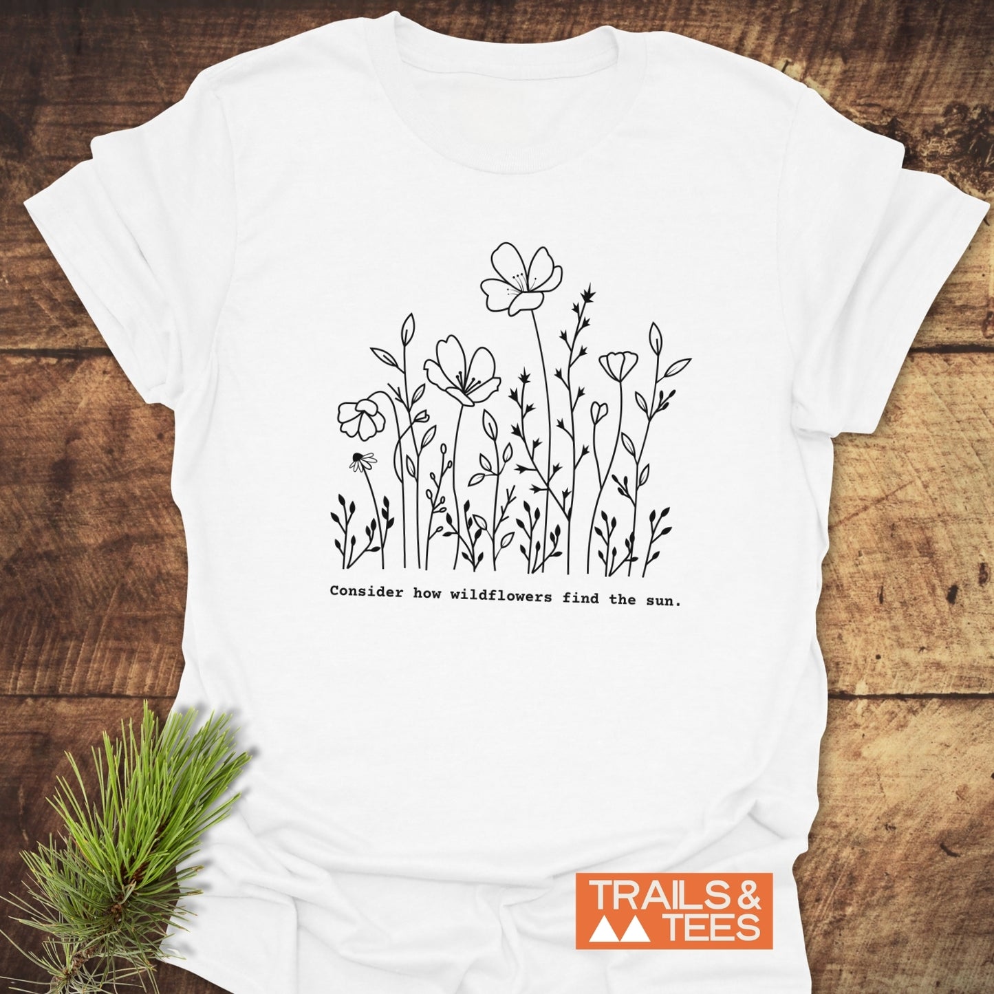 How Wild Flowers Find The Sun T-Shirt: White tee with black wildflower sketch and phrase, “Consider how wildflowers find the sun,” laid flat with Trails & Tees logo, styled with a pine sprig.