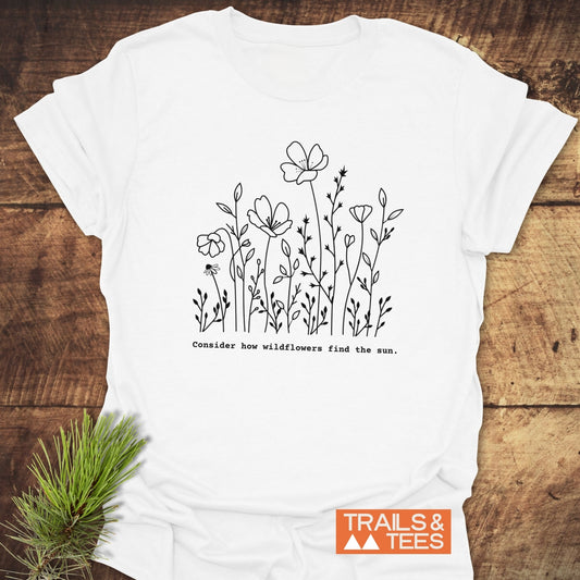 How Wild Flowers Find The Sun T-Shirt: White tee with black wildflower sketch and phrase, “Consider how wildflowers find the sun,” laid flat with Trails & Tees logo, styled with a pine sprig.