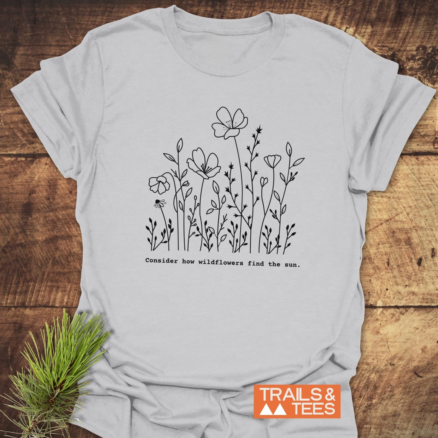 How Wild Flowers Find The Sun T-Shirt features a black wildflower line drawing with a motivational phrase, laid on wood. Includes orange TRAILS & TEES logo, ideal for nature lovers.