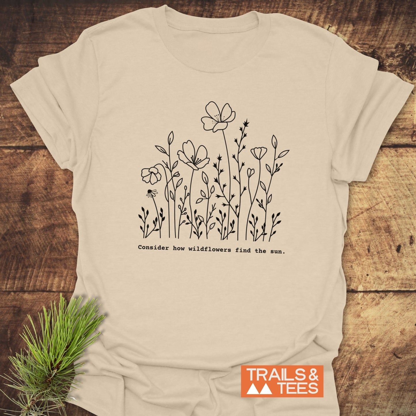 How Wild Flowers Find The Sun T-Shirt: Light-cream tee with black wildflower illustration and phrase, featuring TRAILS & TEES logo. Pine sprig accentuates nature-themed design. Perfect for wildflower enthusiasts.