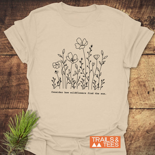 How Wild Flowers Find The Sun T-Shirt: Light-cream tee with black wildflower illustration and phrase, featuring TRAILS & TEES logo. Pine sprig accentuates nature-themed design. Perfect for wildflower enthusiasts.