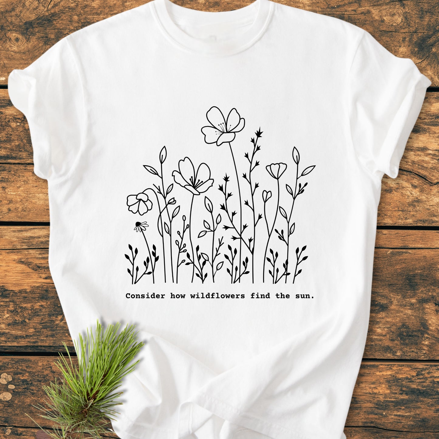 How Wildflowers Find The Sun - Vintage Wash Tee: A flat-lay of a white T-shirt featuring black line-drawn wildflowers and a typewriter-style quote, with a pine sprig accentuating the natural theme.