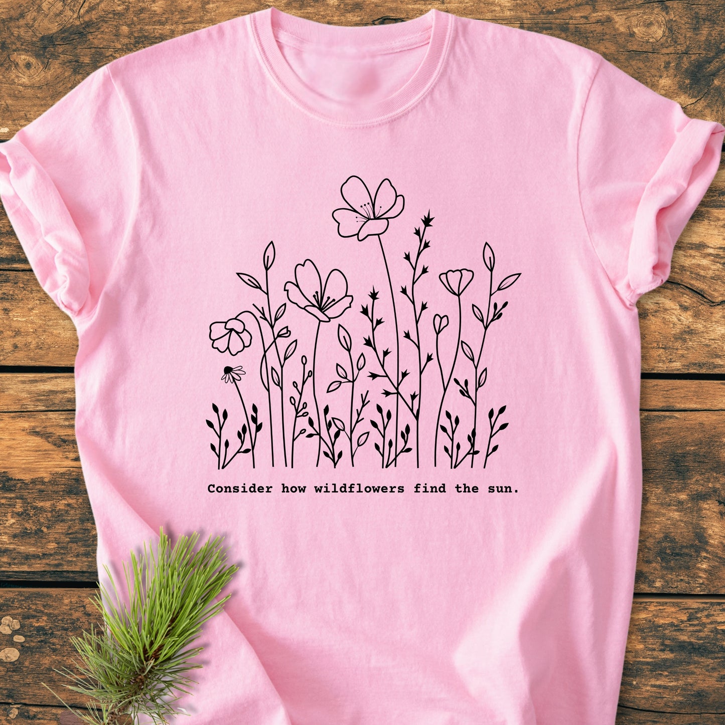 How Wildflowers Find The Sun - Vintage Wash Tee featuring a black line-drawing of wildflowers and phrase on a soft-pink fabric, laid on wood with pine needles, embodies nature-inspired relaxation.