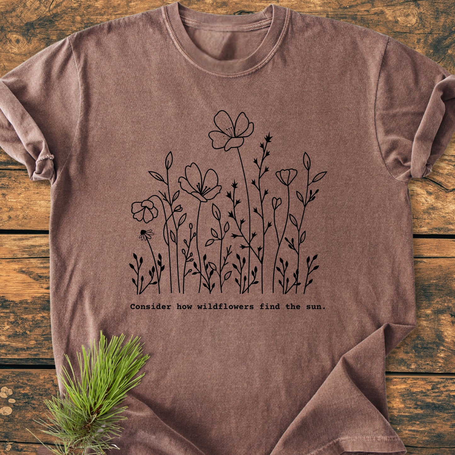 How Wildflowers Find The Sun - Vintage Wash Tee features a line-drawing of wildflowers with an inspirational quote, laid flat on wood, accented by a small pine sprig for natural styling.