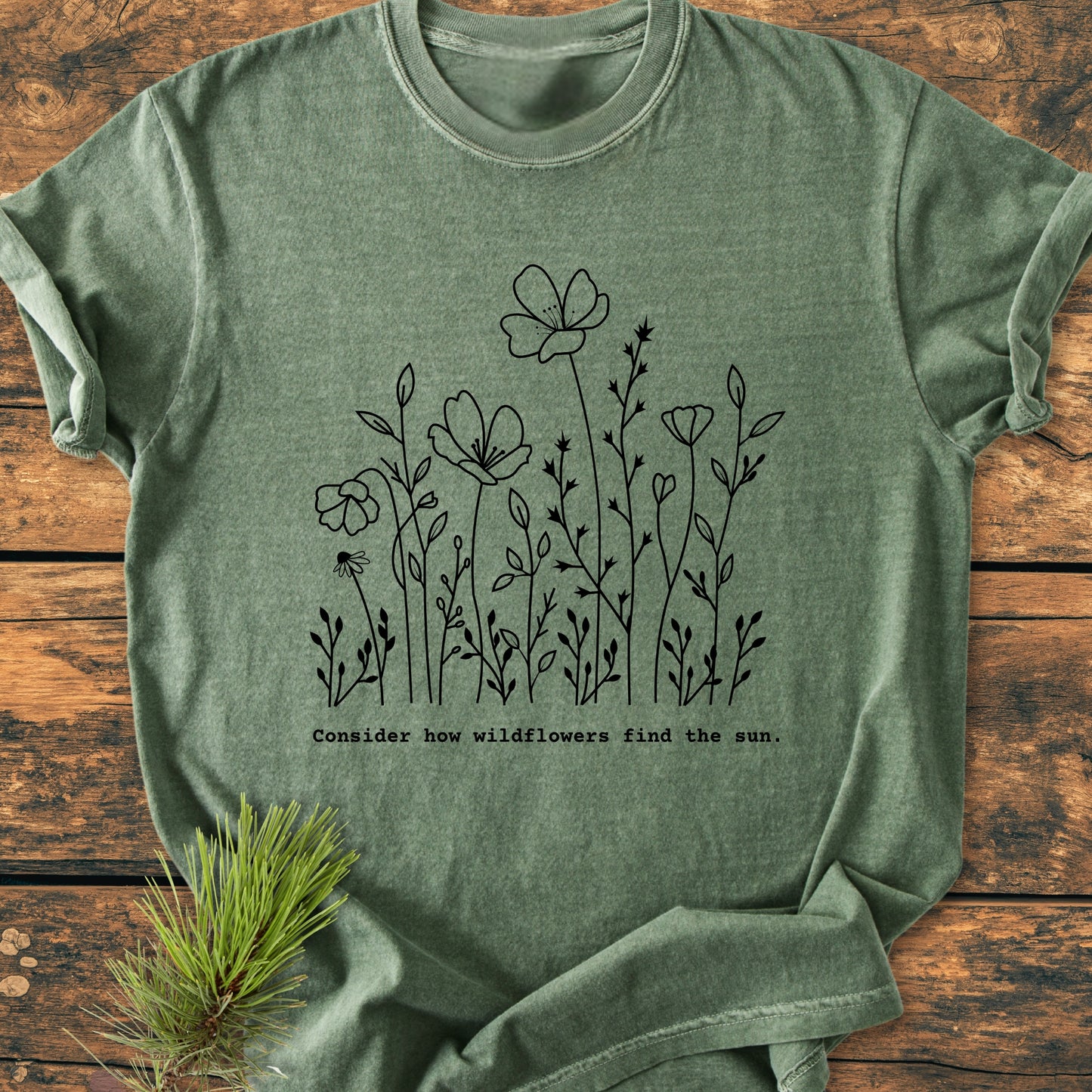 How Wildflowers Find The Sun - Vintage Wash Tee, featuring a black line-drawing of wildflowers and text, lies flat with pine needles beside it, embodying a nature-inspired theme on a wooden surface.