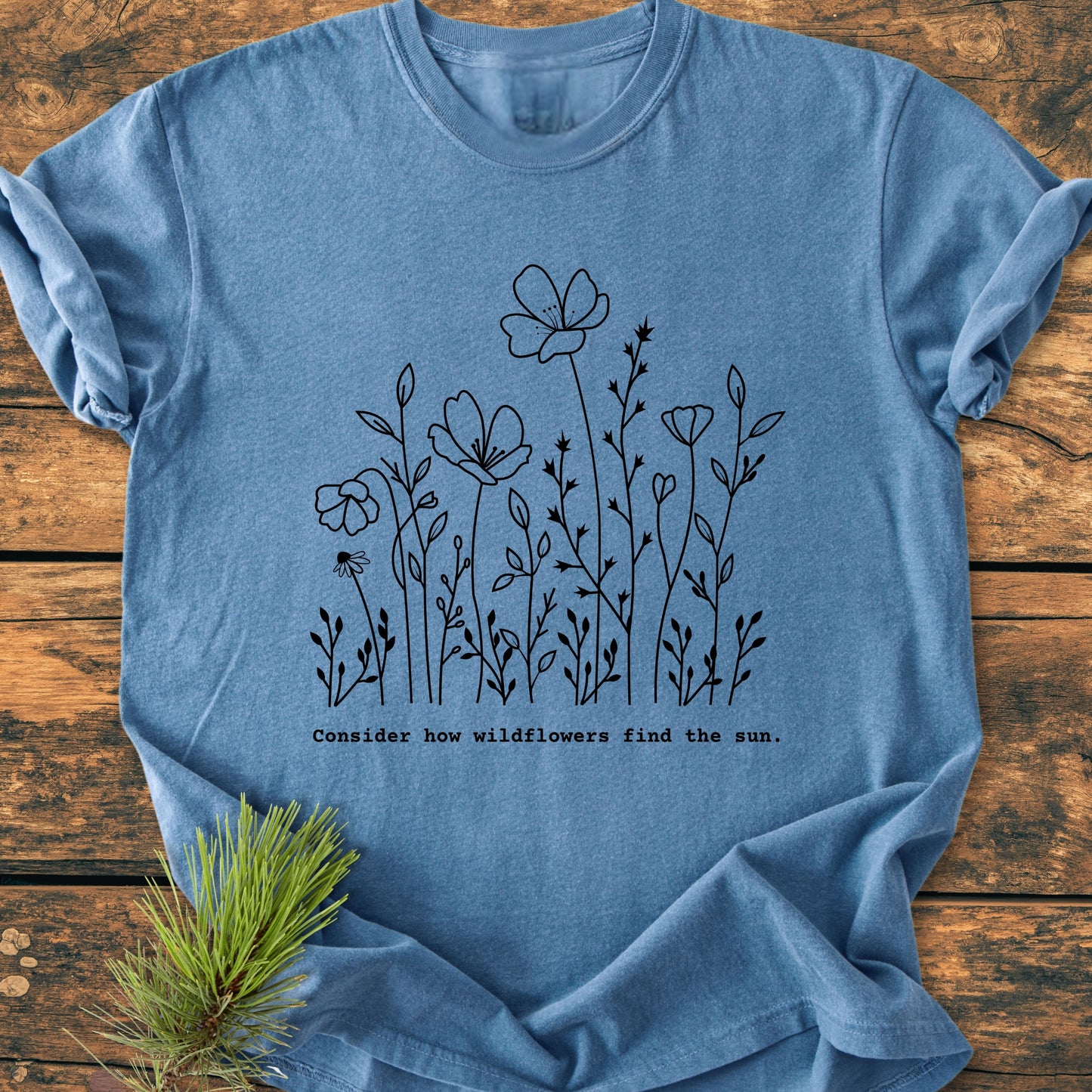 How Wildflowers Find The Sun - Vintage Wash Tee features a black wildflower sketch and uplifting phrase, displayed with a pine sprig, highlighting its botanical theme and rugged charm.