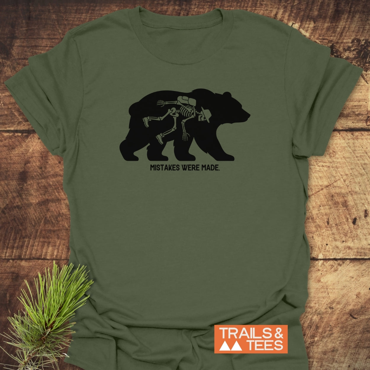 Mistakes Were Made T-Shirt with a bear silhouette featuring a hiker skeleton, captioned below. Laid on a wooden surface with pine sprig, includes TRAILS & TEES logo in the corner.
