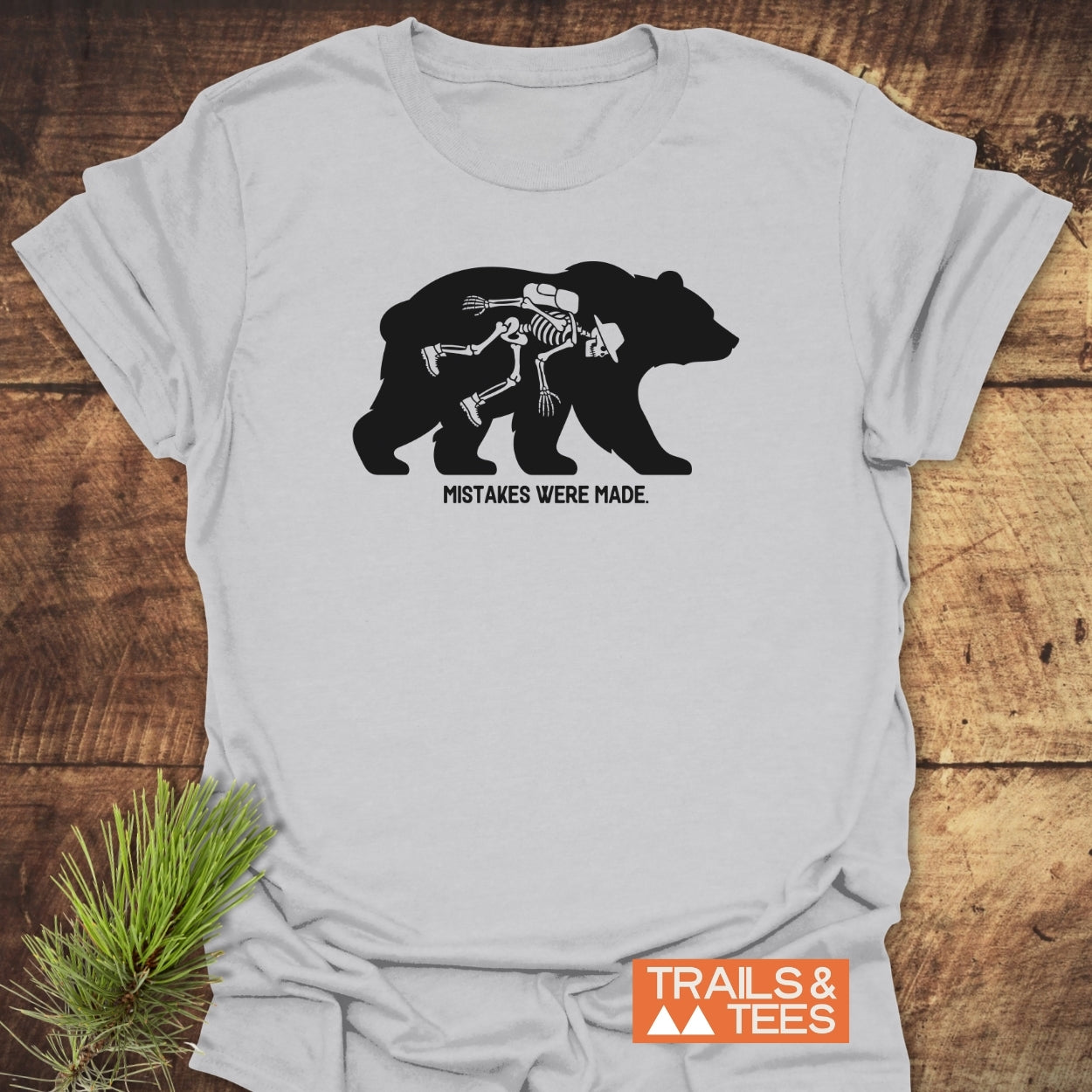 Mistakes Were Made T-Shirt featuring a bear silhouette with a skeleton hiker inside, captioned below. Displayed on a wooden surface with a pine sprig, emphasizing dark trail humor.