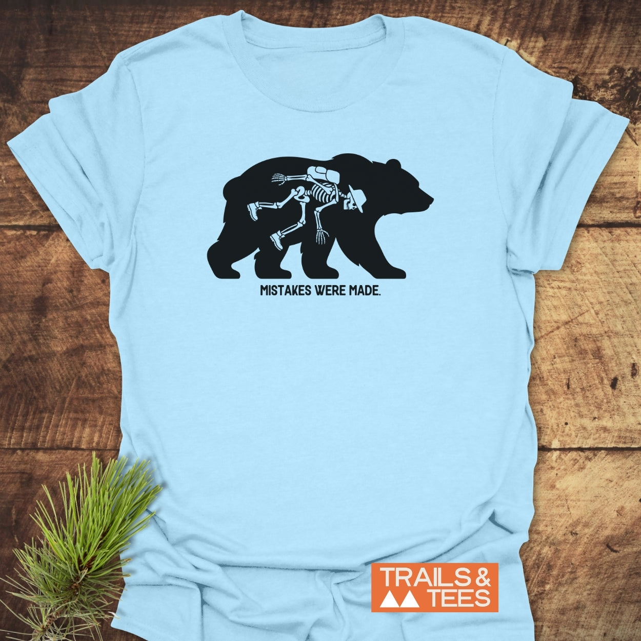 Light-blue Mistakes Were Made T-Shirt features a black bear silhouette with a cartoon skeleton hiker inside, captioned Mistakes Were Made, and Trails & Tees logo in the corner.