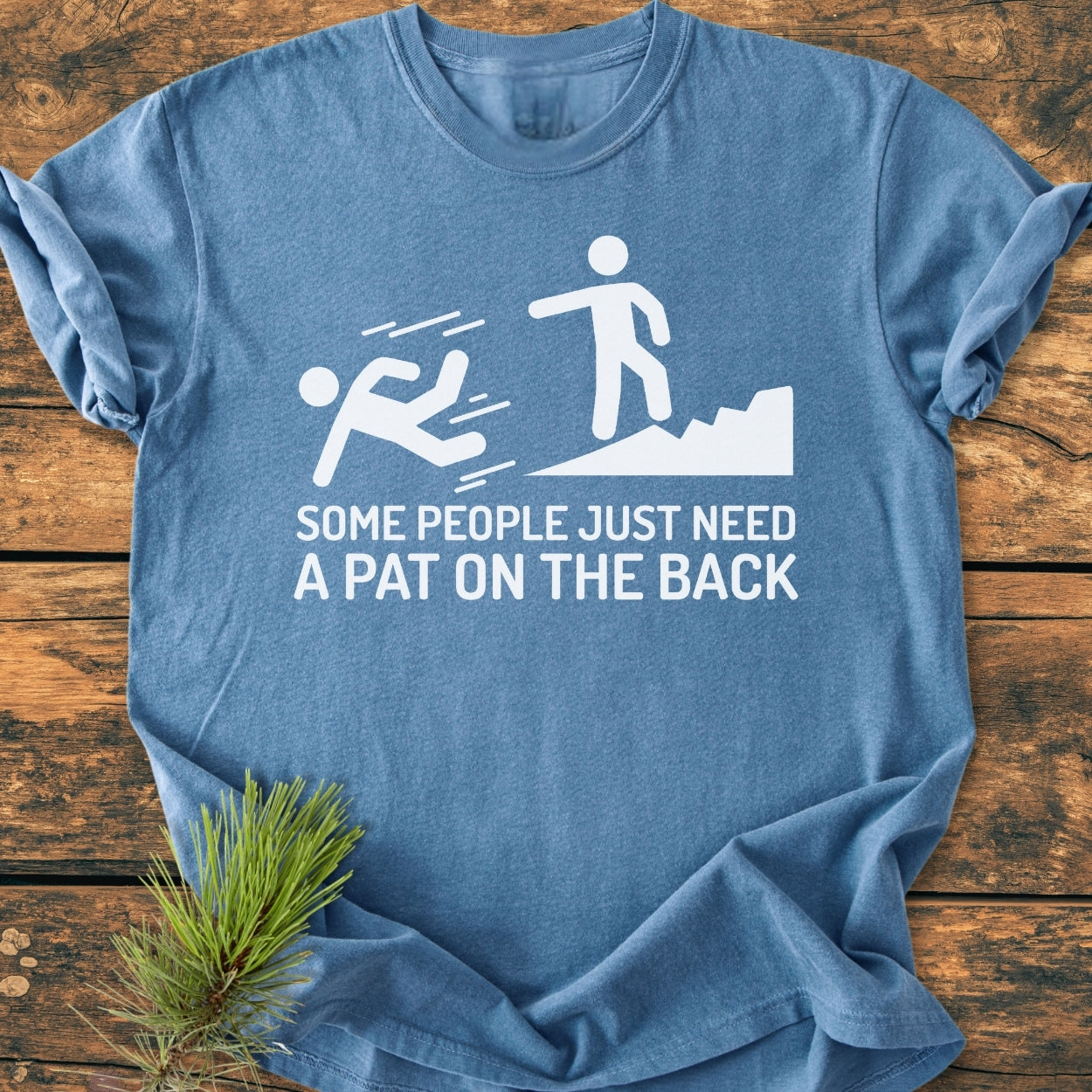 Some People Just Need A Pat On The Back - Vintage Wash Tee featuring a playful stick-figure cartoon with a caption, laid on a table with a pine branch, highlighting its hiking theme.