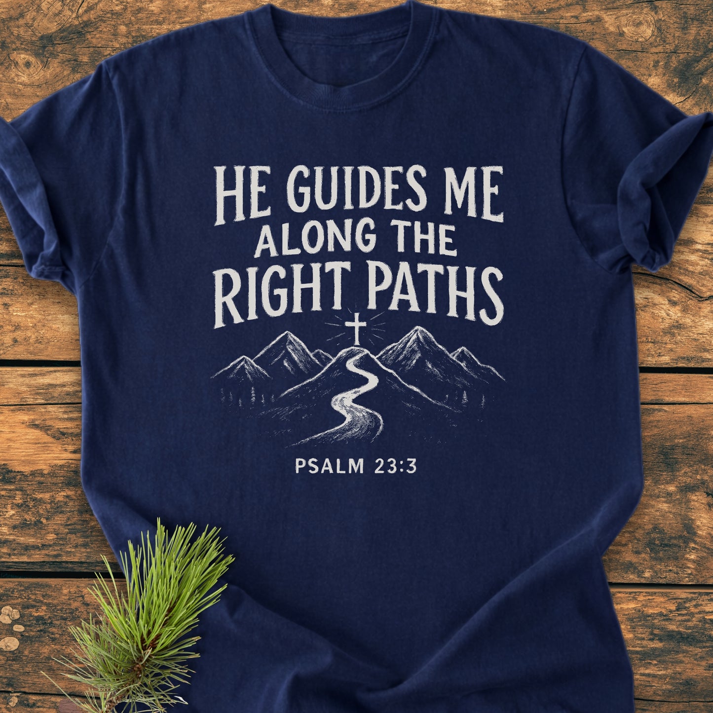 He Guides Me - Vintage Wash Tee featuring a mountain and trail sketch with Psalm 23:3, displayed on a wooden background, complemented by a pine sprig, symbolizing faith and guidance.