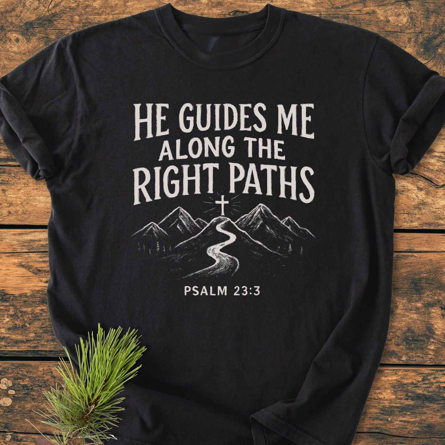He Guides Me - Vintage Wash Tee, featuring a hand-drawn mountain path design with scripture, laid on wood with pine sprig; embodies faith, guidance, and nature's tranquility.