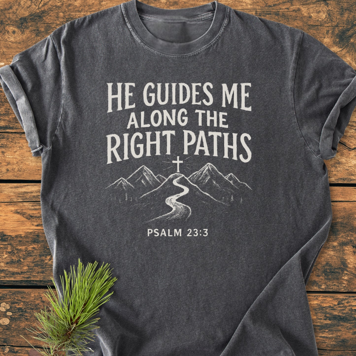 He Guides Me - Vintage Wash Tee featuring a winding mountain road graphic with a cross above and Psalm 23:3 text, laid out with a pine sprig on a wooden background.