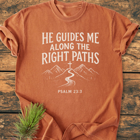 He Guides Me - Vintage Wash Tee: Rust-orange cotton shirt with a white mountain path graphic and Psalm 23:3 quote, laid on wood with pine sprig; symbolizes divine guidance for Christian hikers.