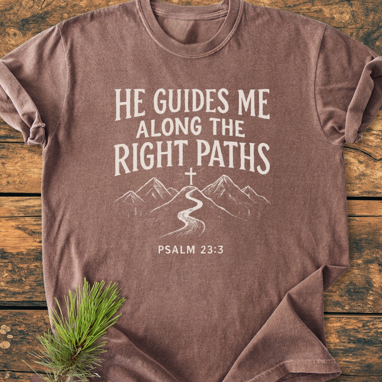 He Guides Me - Vintage Wash Tee featuring Psalm 23:3 with a mountain path design, laid on rustic wood with pine. A soft, relaxed fit shirt symbolizing faith and divine guidance.