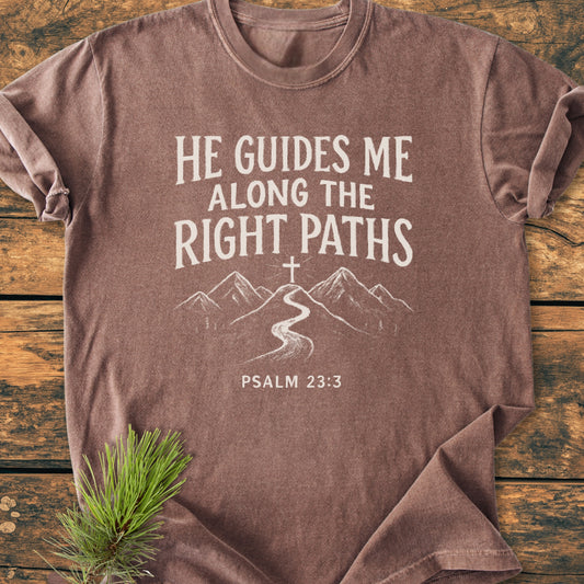 He Guides Me - Vintage Wash Tee featuring Psalm 23:3 with a mountain path design, laid on rustic wood with pine. A soft, relaxed fit shirt symbolizing faith and divine guidance.