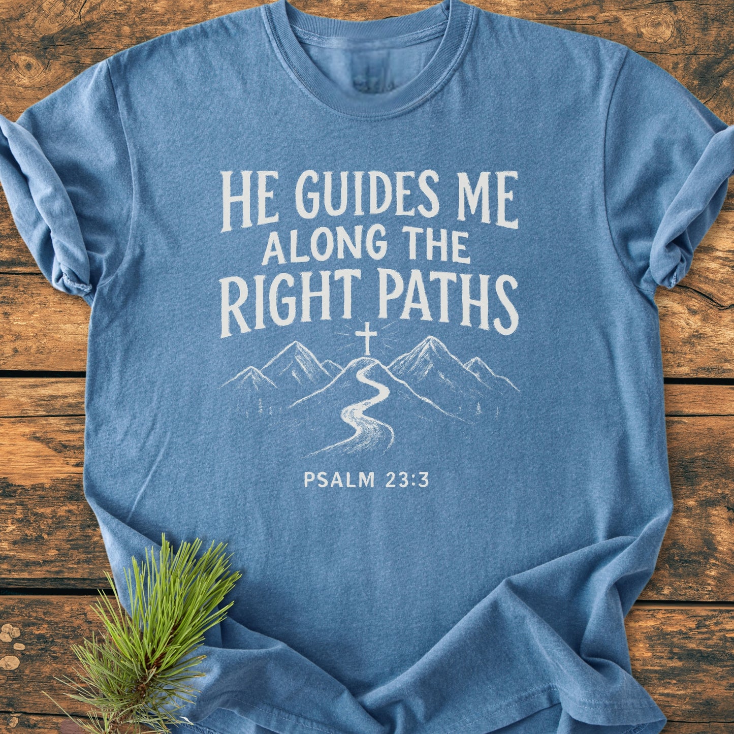 Light-blue He Guides Me - Vintage Wash Tee with a white design of Psalm 23:3, featuring a stylized cross and mountain trail, laid on a wooden surface with pine sprig.