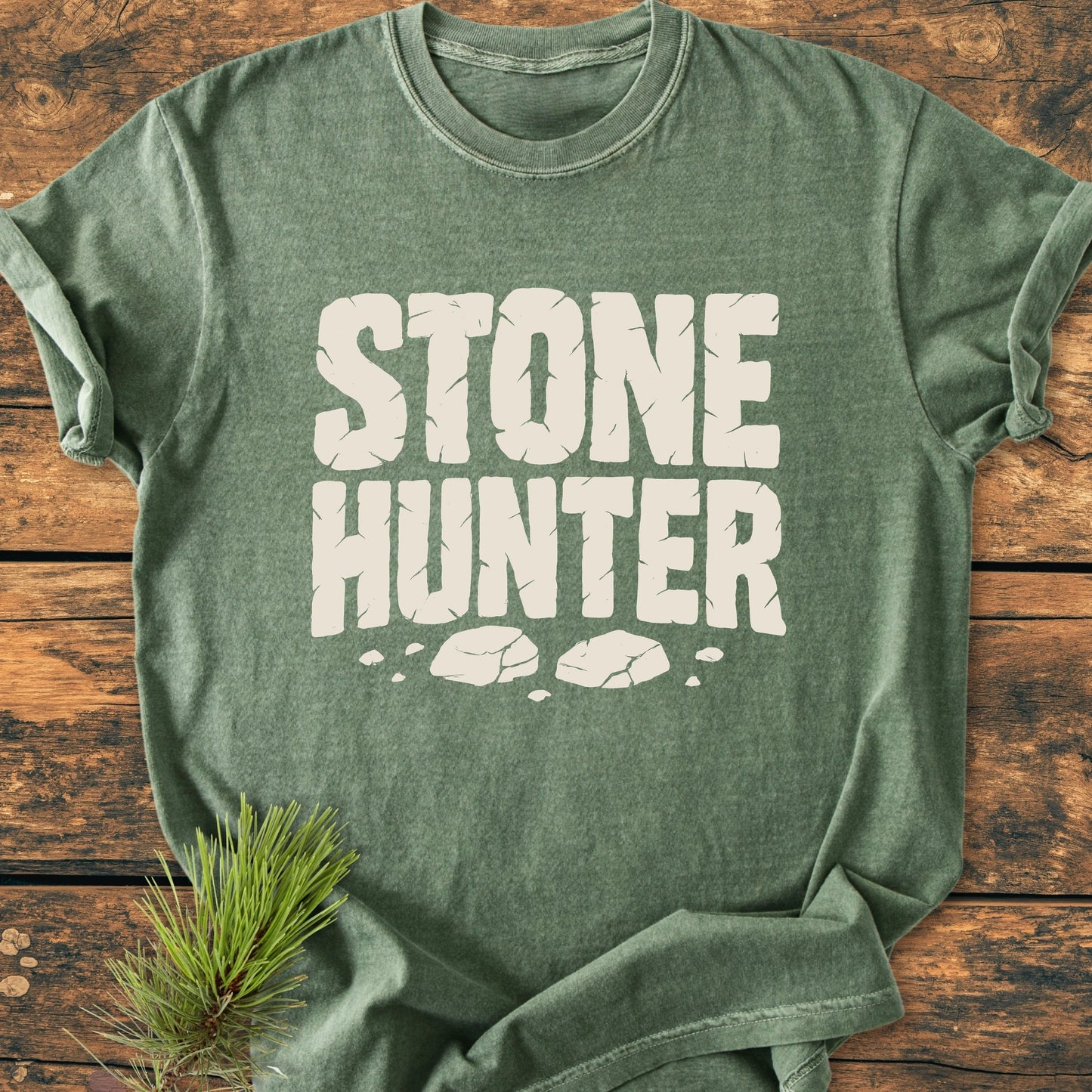 Heather-green STONE HUNTER tee with white text and cracked stone graphics on rustic wood. Vintage wash for a soft, broken-in feel. Pine branch accentuates outdoor adventure theme.