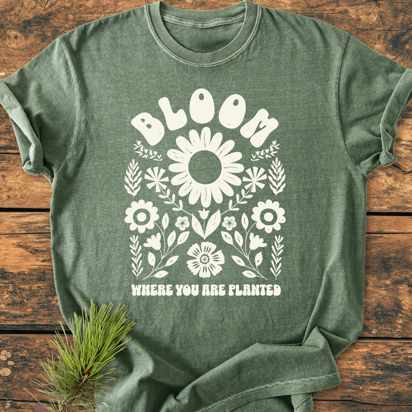 Olive-green Bloom Where You Are Planted T-shirt featuring a floral graphic, highlighting a large blossom and leafy sprigs, laid flat on wood with a pine sprig beside it.