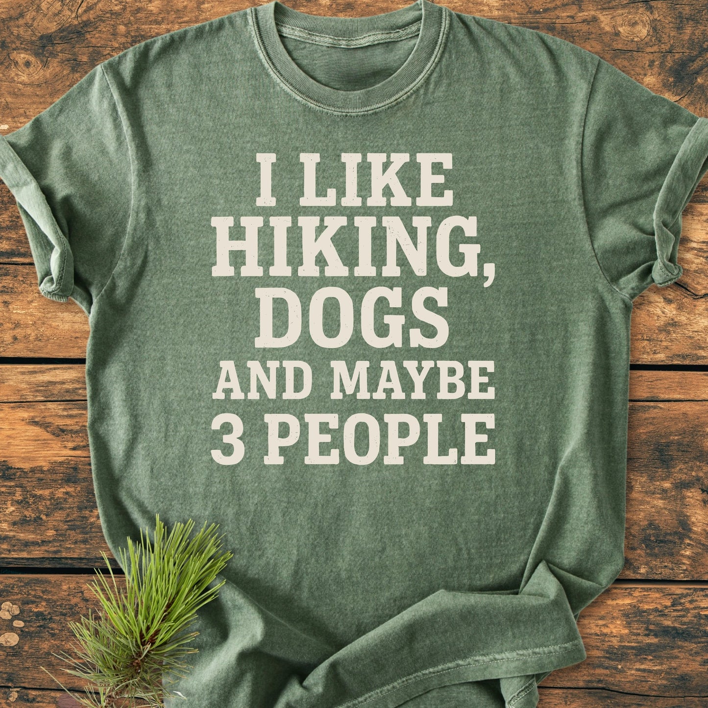 Folded green 1431 I Like Hiking And Dogs T-shirt on wood, featuring bold white slogan. Accented by pine sprig, showcasing its vintage wash and relaxed fit, perfect for outdoor enthusiasts.