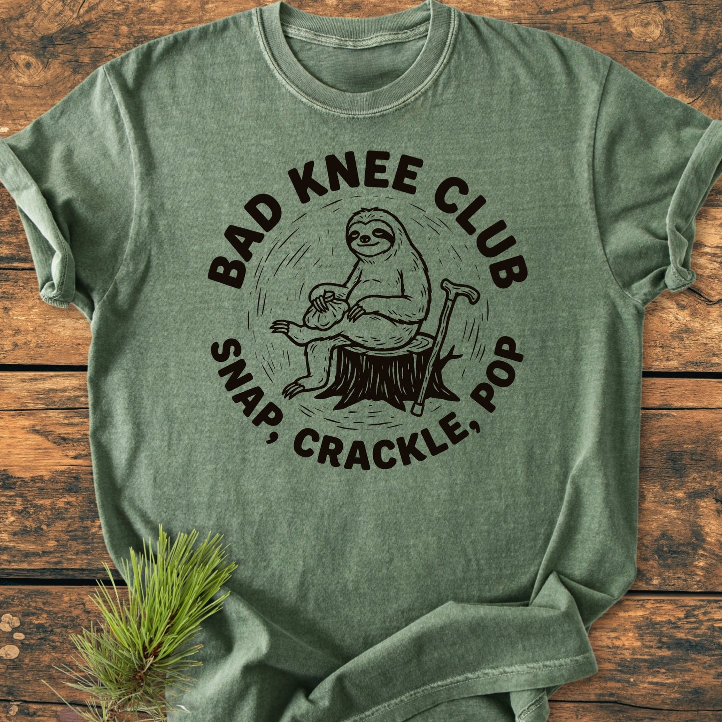 Heather-green T-shirt featuring BAD KNEE CLUB graphic with a sloth on a stump and SNAP, CRACKLE, POP slogan, styled with a pine sprig for a vintage, relaxed look.