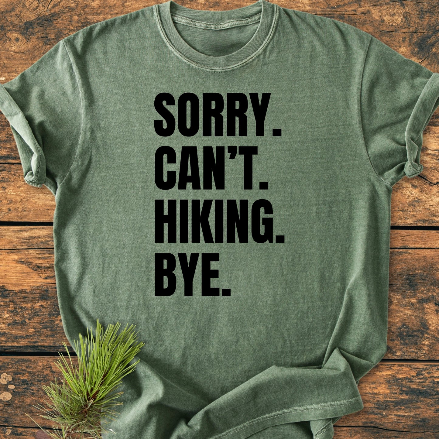 Heather-green T-shirt with SORRY. CAN’T. HIKING. BYE. slogan in bold letters, laid flat on a rustic wooden surface with a pine sprig nearby, showcasing a vintage wash and relaxed fit.