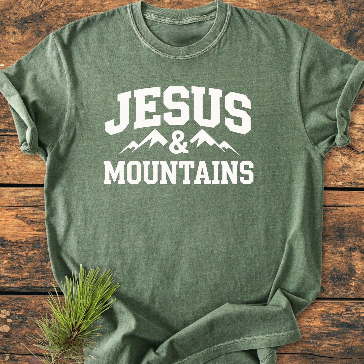 Heather-green crew-neck T-shirt with “JESUS & MOUNTAINS” in white, featuring a mountain graphic, laid on rustic wood. Ideal for a relaxed, vintage style and everyday comfort.