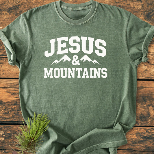 Heather-green crew-neck T-shirt with “JESUS & MOUNTAINS” in white, featuring a mountain graphic, laid on rustic wood. Ideal for a relaxed, vintage style and everyday comfort.