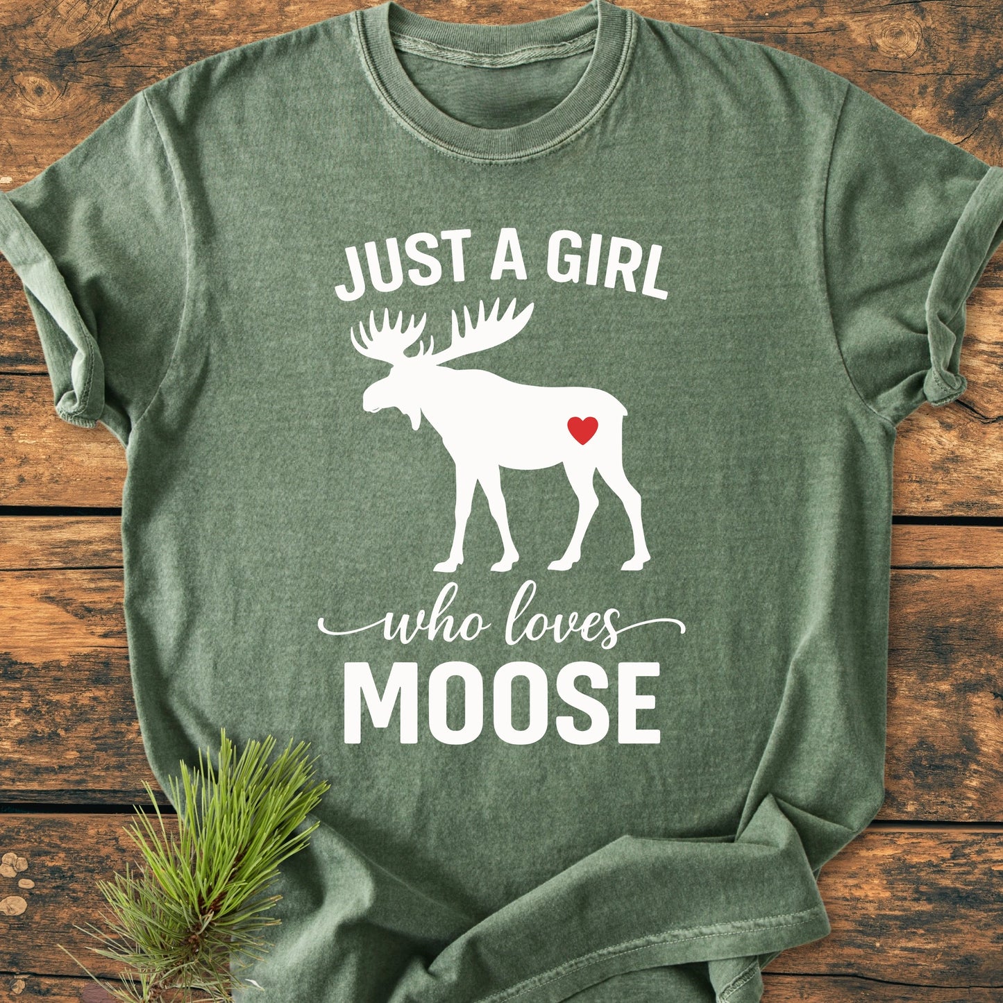 Green T-shirt, 1437 Just A Girl Who Loves Moose, features white moose silhouette with red heart, white text. Laid on wood, pine sprig nearby. Soft, relaxed fit, vintage wash style.