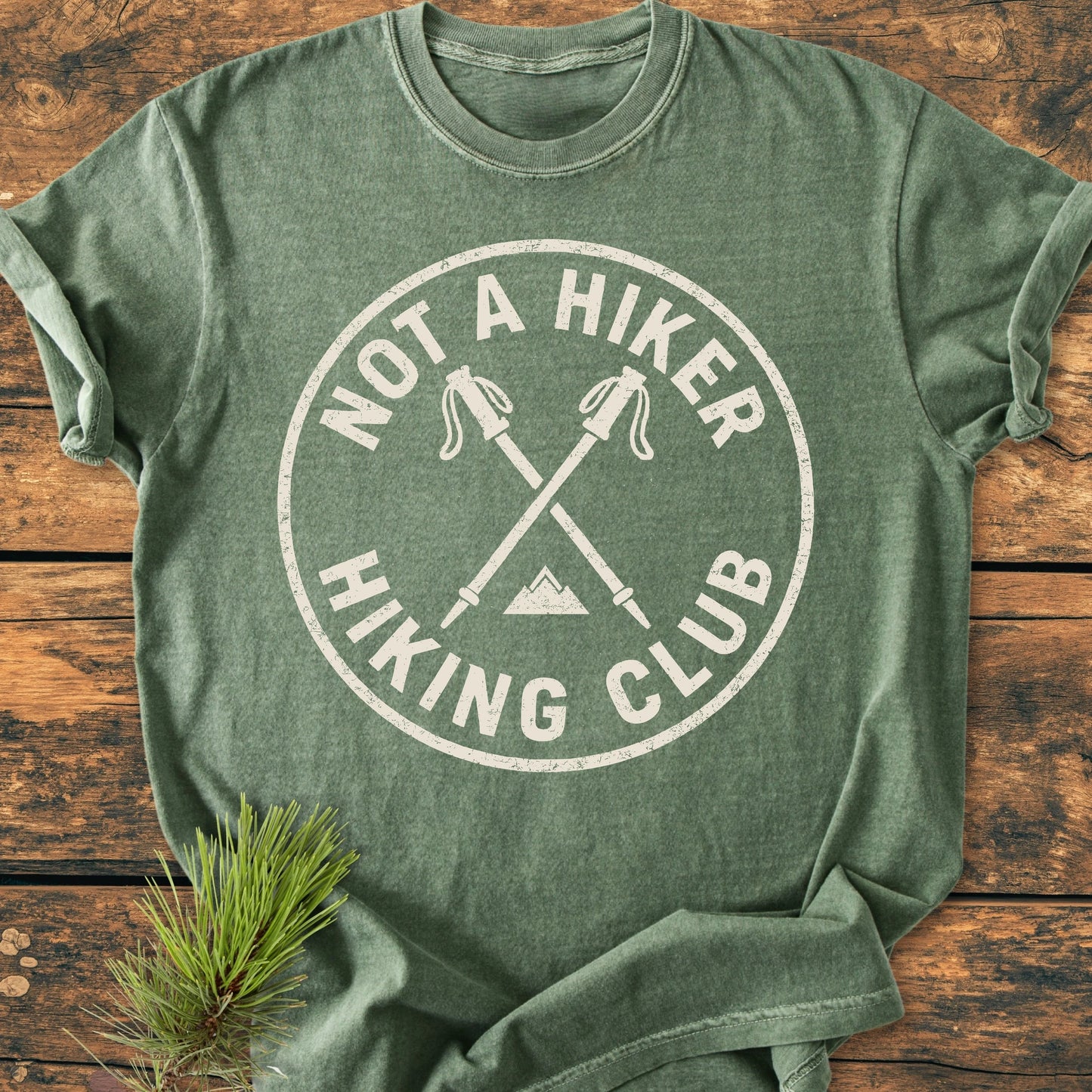 Heather-green T-shirt with NOT A HIKER Hiking Club logo, featuring crossed poles and mountain peak, laid flat with pine sprig. Vintage wash for everyday wear, relaxed fit for comfort.