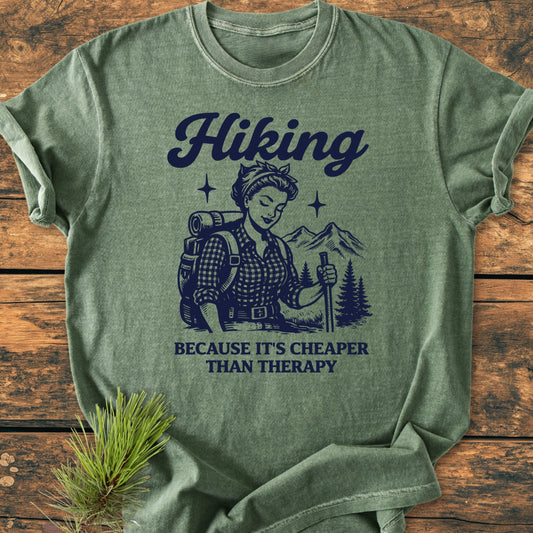 1407 Hiking Cheaper Than Therapy Base T-shirt, featuring a vintage hiker illustration with pine trees and mountains, folded on wood. Includes “Hiking” and slogan, emphasizing comfort and durability.