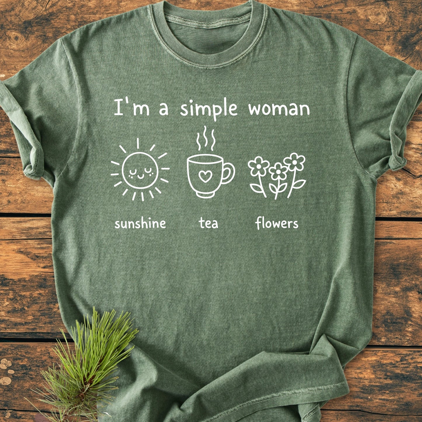 1408 I'm A Simple Woman T-shirt, moss-green, features a smiling sun, steaming cup, and flowers. Crafted from 100% ring-spun cotton for comfort and durability, laid on a wooden surface.