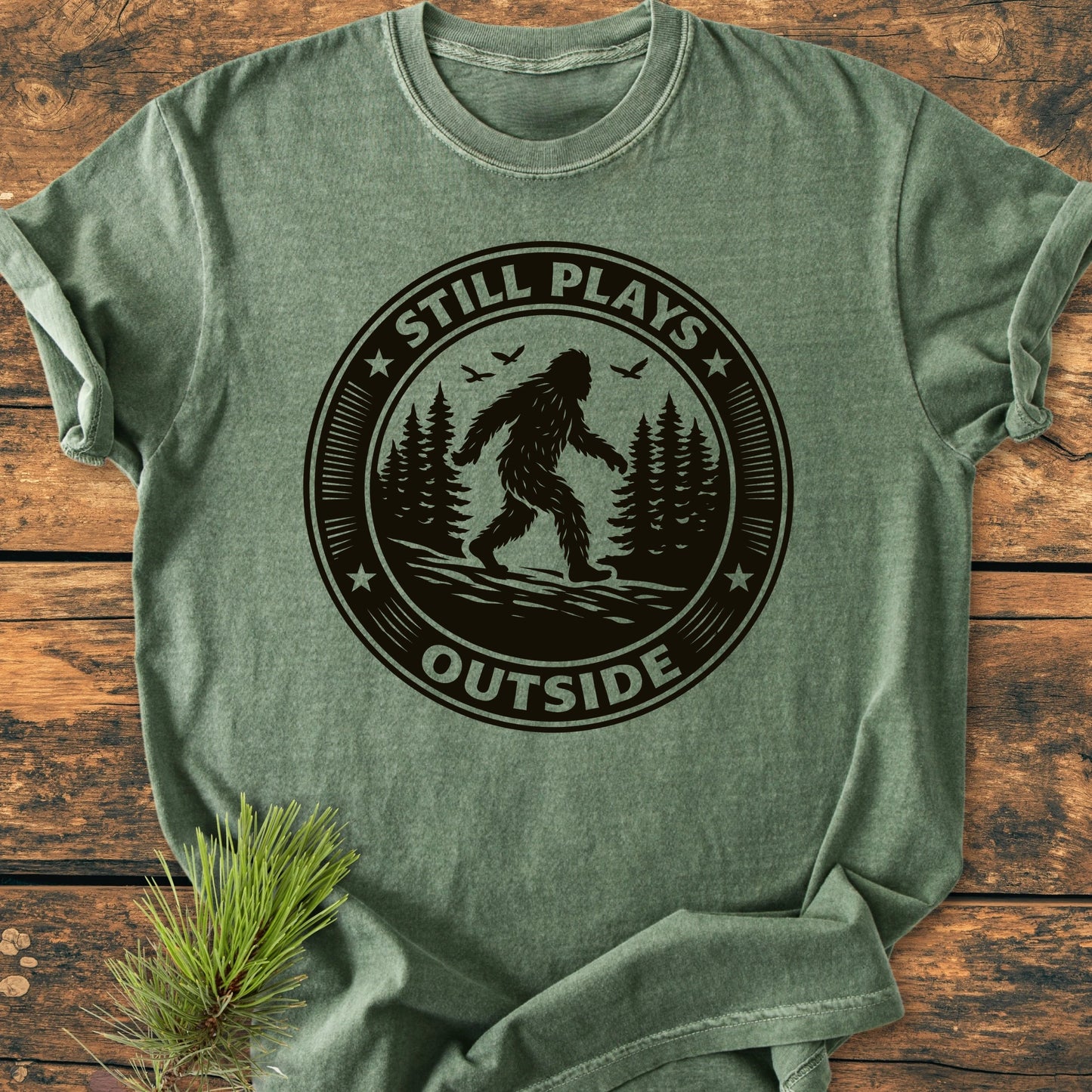 Green T-shirt with 1430 Still Plays Outside Bigfoot design, featuring a Sasquatch silhouette and pine trees. Laid on wood, includes a pine sprig. Perfect for outdoor enthusiasts, vintage wash fabric.