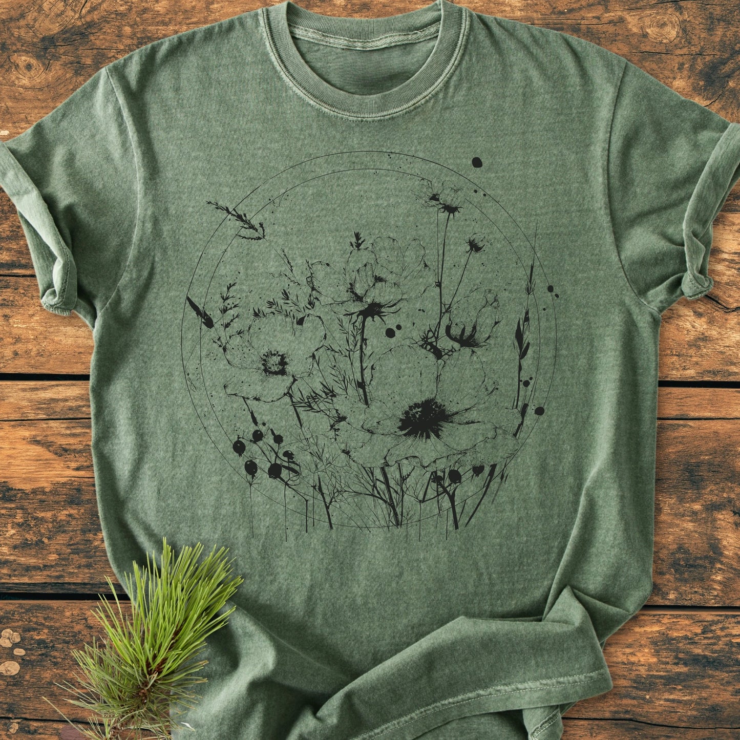 The 391 Wildflower Circle T-shirt features a black line-art print of poppies on a heather-green fabric, laid on wood, showcasing its relaxed fit and soft texture.