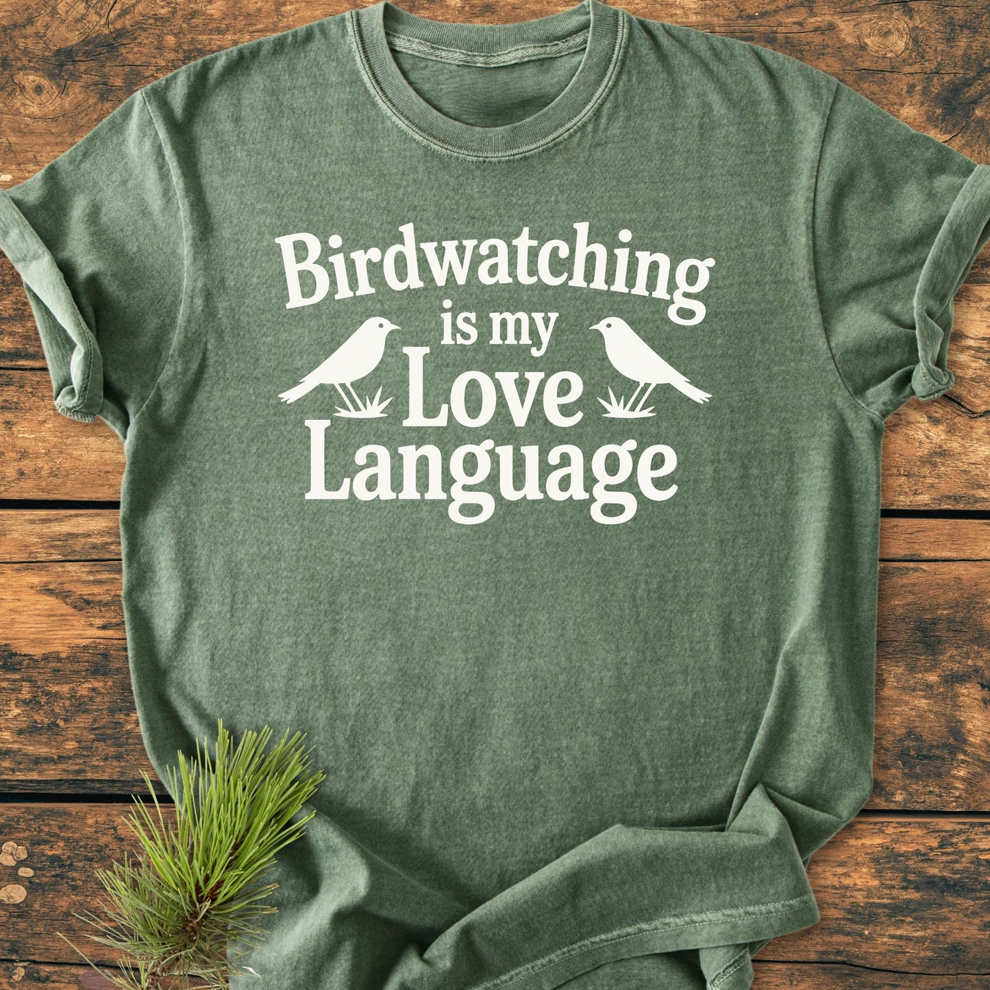 Forest-green T-shirt titled 1428 Birdwatching Love Language features white lettering and bird silhouettes, laid flat on wood. Includes vintage wash, relaxed fit, and durability for everyday wear.