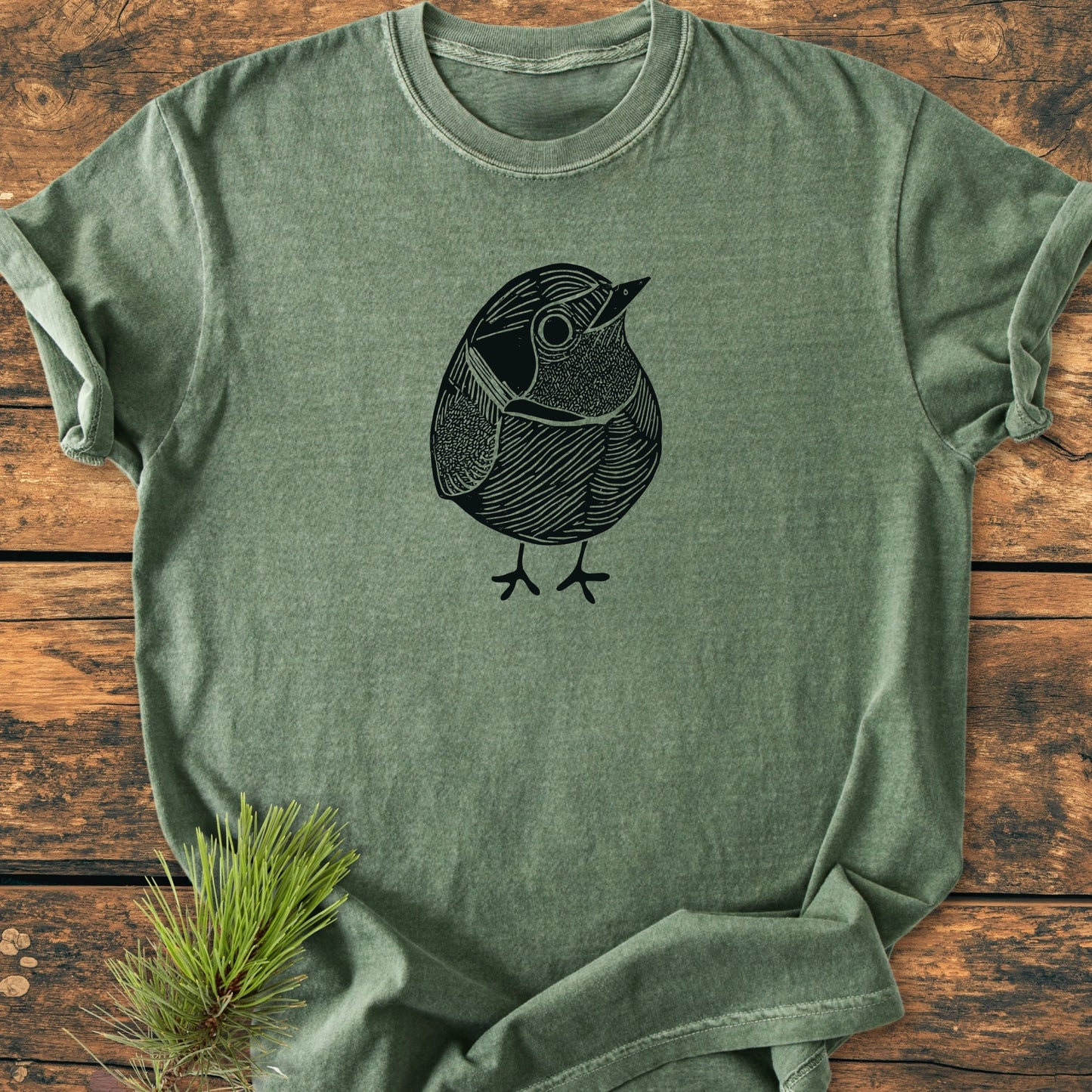 Heather-green crew-neck T-shirt titled 1433 Little Robin, featuring a black line-art bird illustration, laid flat on wood with a pine sprig for a vintage, relaxed style.