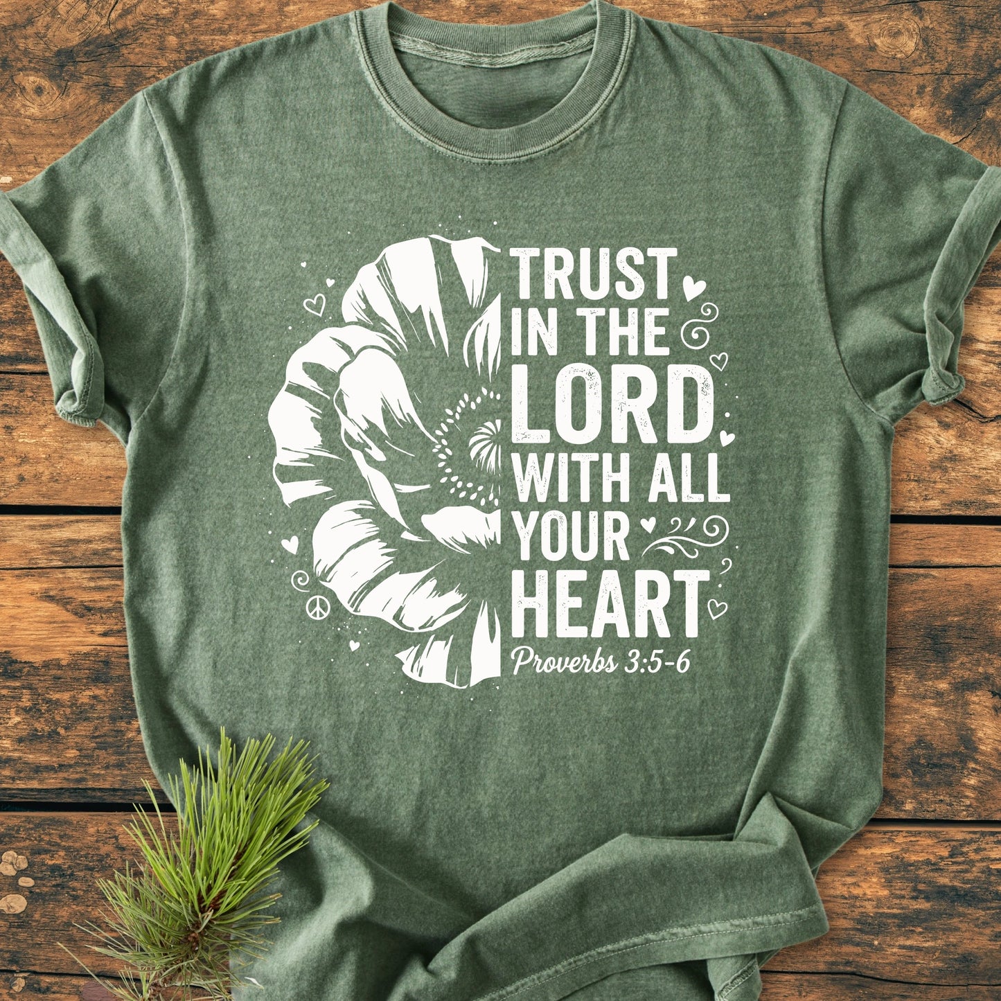 Trust In The Lord - Vintage Wash Tee, featuring a large stylized flower and Proverbs 3:5–6, laid flat with pine sprig on wood, showcasing its relaxed, soft-washed, unisex fit.