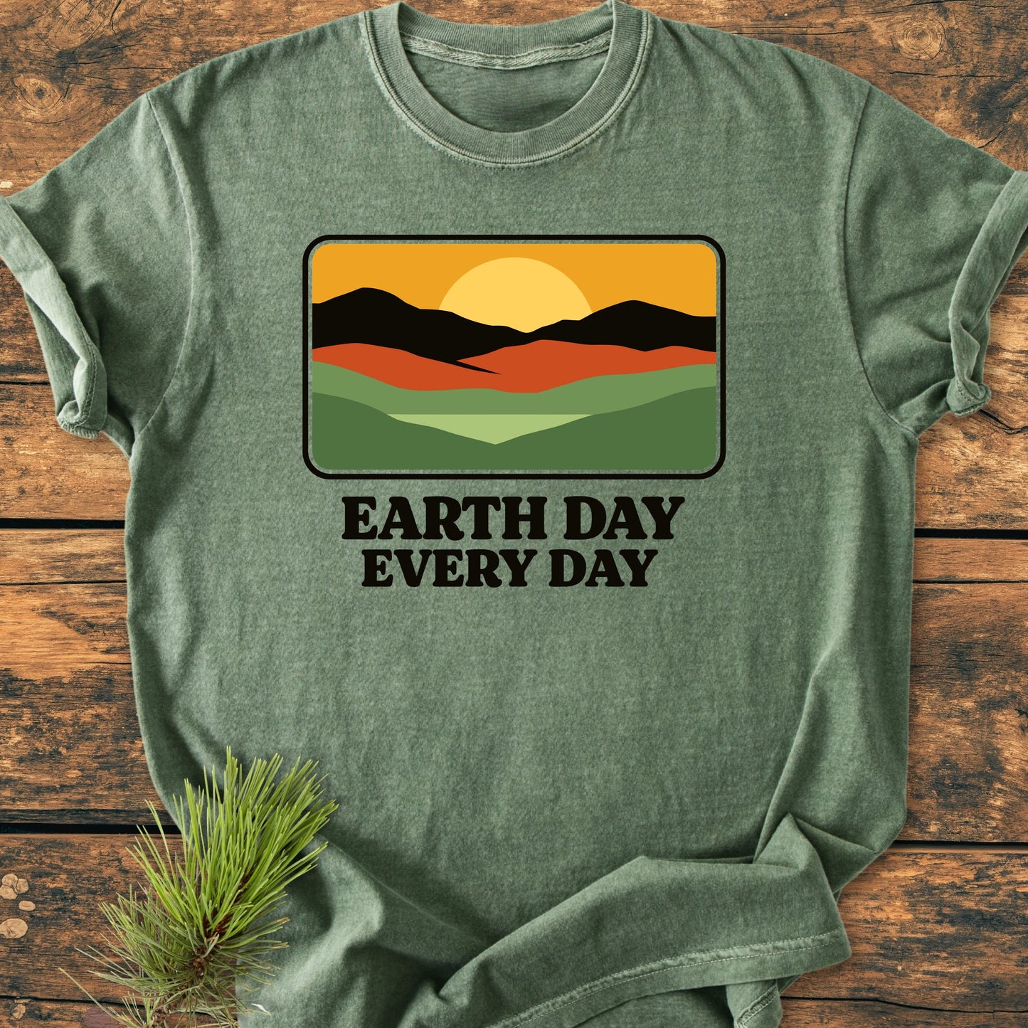 Green T-shirt with EARTH DAY EVERY DAY graphic of hills and sun, laid on wood. Vintage wash for a relaxed, soft-washed fit, ideal for everyday adventures.