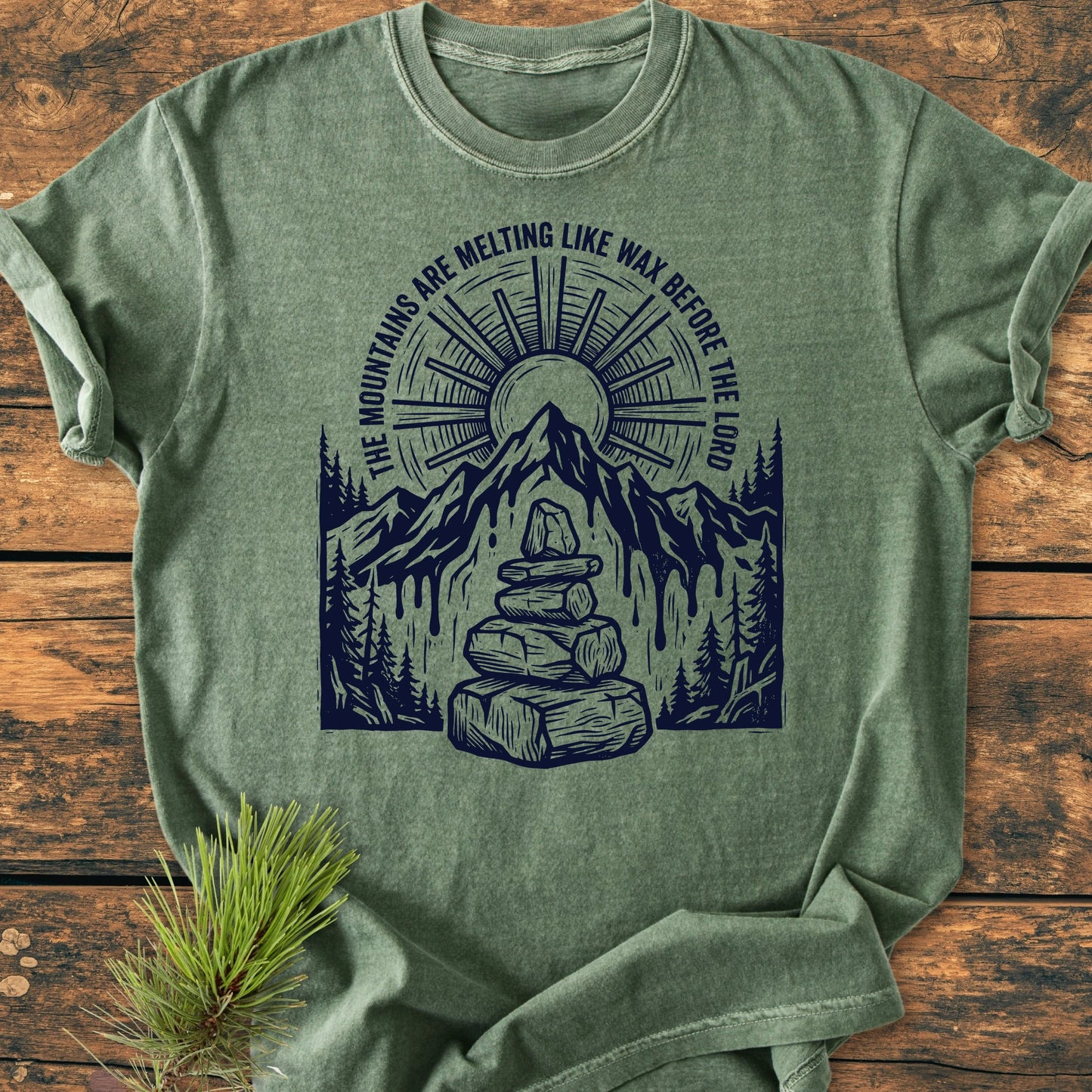 Heather-green T-shirt, titled 1414 Mountains Melting Like Wax, features a stylized outdoor scene with cairn, pine trees, mountain peak, and sunburst, accompanied by the phrase above the graphic.