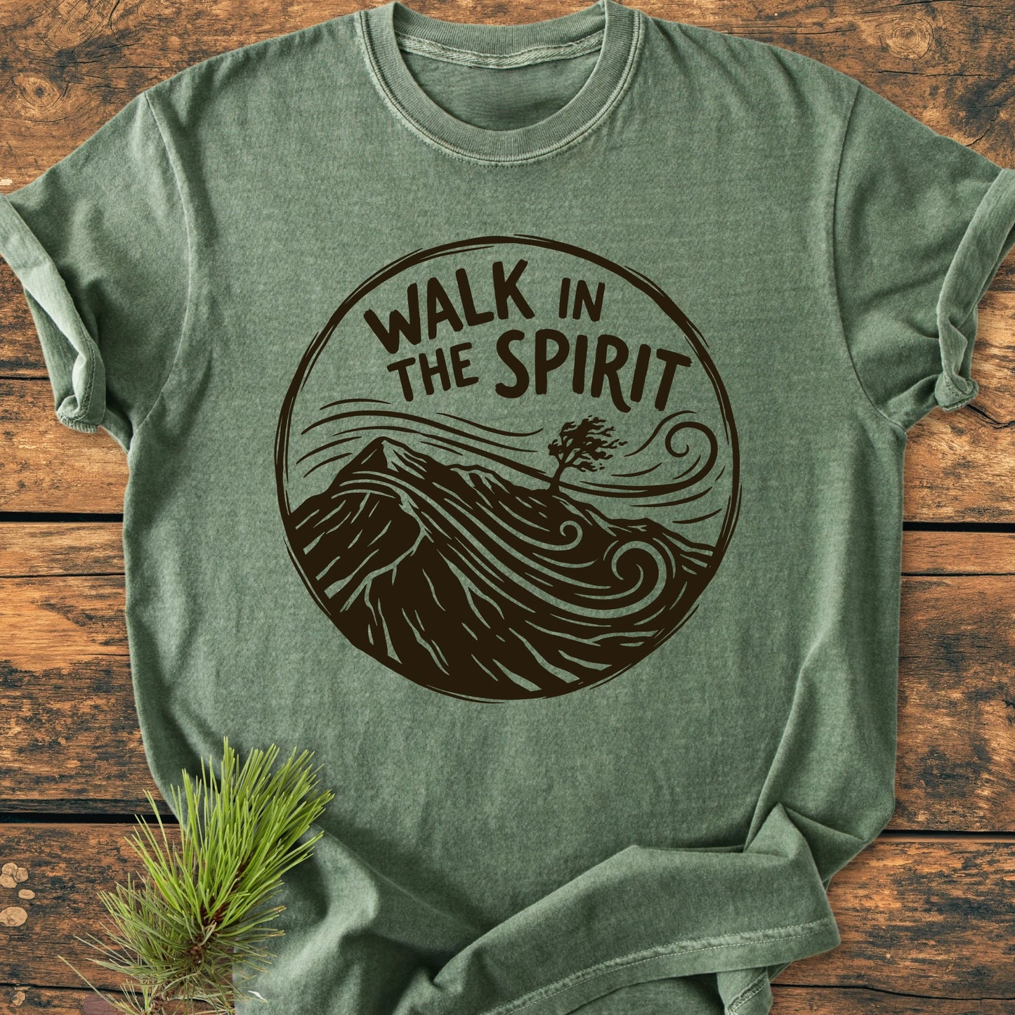 Heather-green 1410 Walk In The Spirit T-shirt featuring a dark-brown graphic with a mountain ridge and tree, laid on wood. Pine sprig nearby, highlighting the shirt's nature-inspired design.
