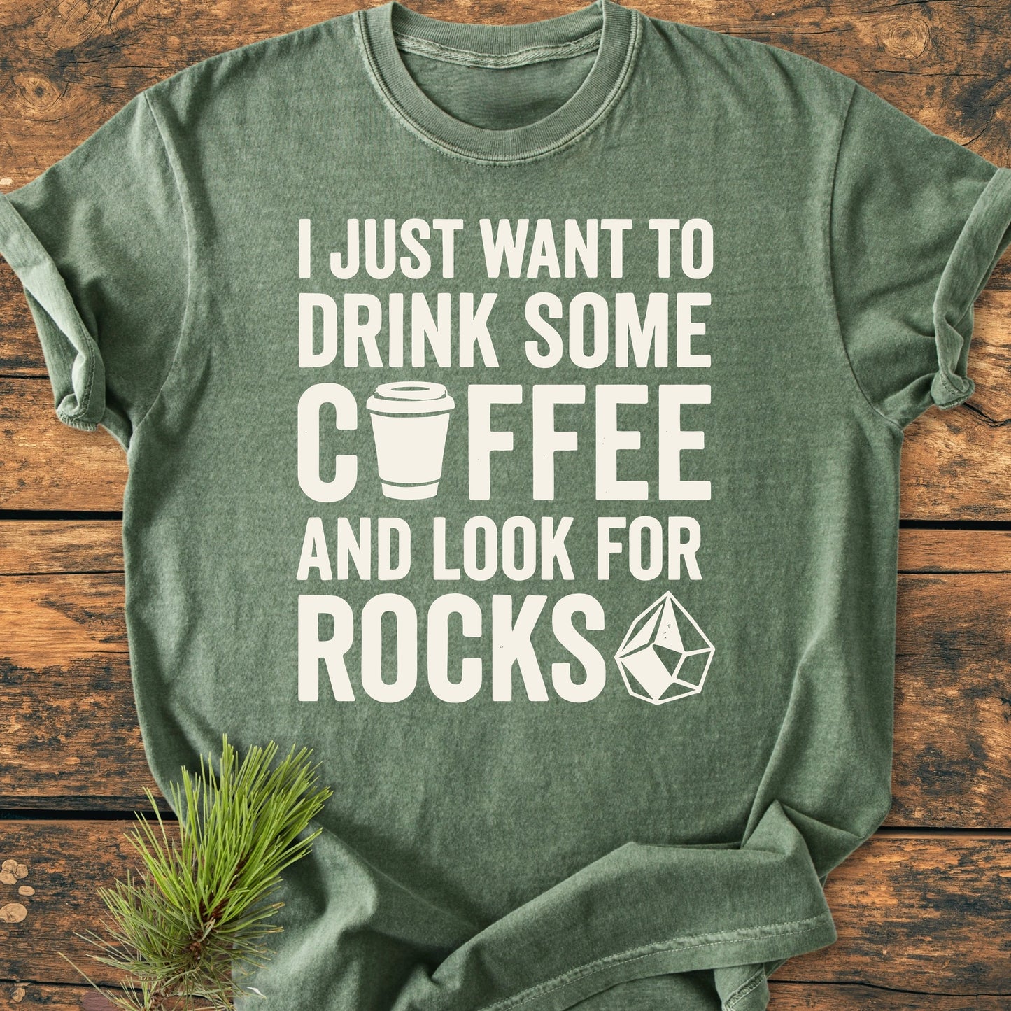 Heather-green 1404 Drink Coffee And Look For Rocks Base T-shirt, features playful white text and graphics, displayed flat with pine sprig. Highlights comfort, ring-spun cotton, and durable double-needle stitching.