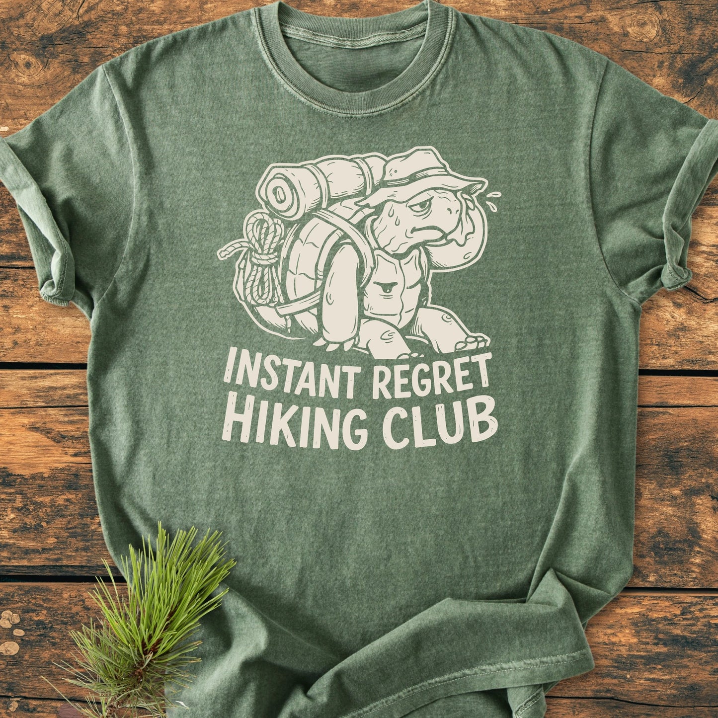 Green T-shirt featuring 1430 Instant Regret Hiking Club with a cartoon turtle wearing a hiking pack and bucket hat, laid on a wooden surface with a pine sprig nearby.