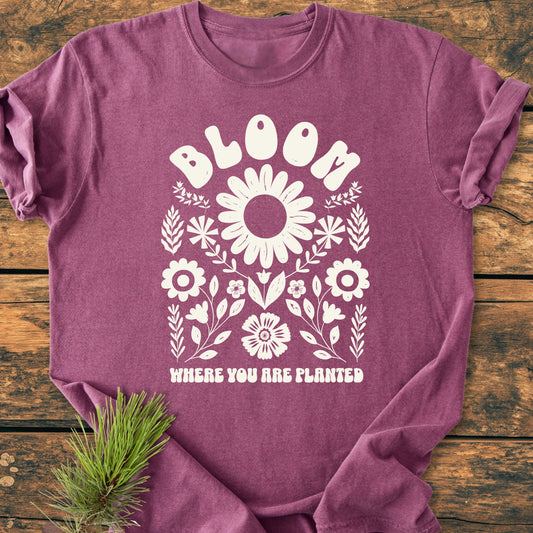 Heather-purple Bloom Where You Are Planted T-shirt features a white folk-art floral design and uplifting text, laid on wood with pine needles. Crafted for a vintage feel and relaxed fit.