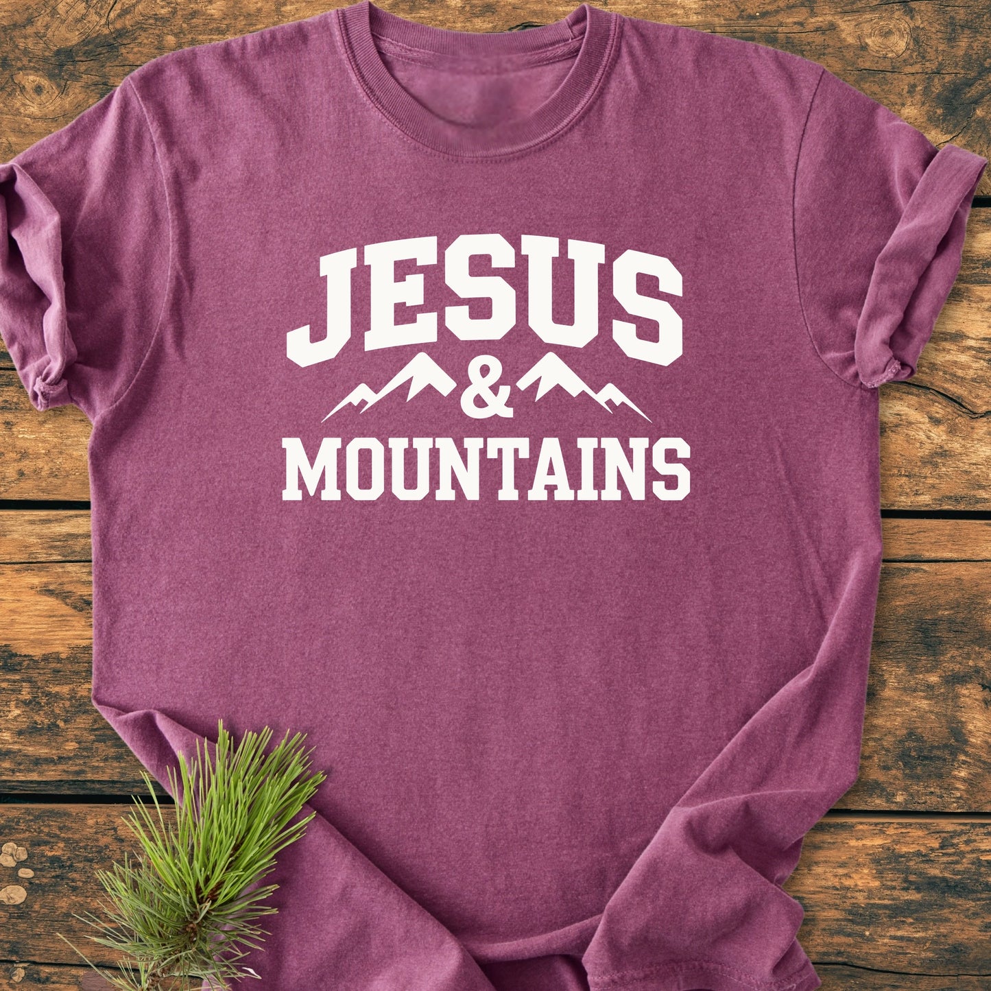 Heathered-purple T-shirt with JESUS & MOUNTAINS and mountain graphic, laid flat on wood. Beside it, a pine twig. Soft-washed, relaxed fit, vintage feel. Perfect for outdoor enthusiasts.